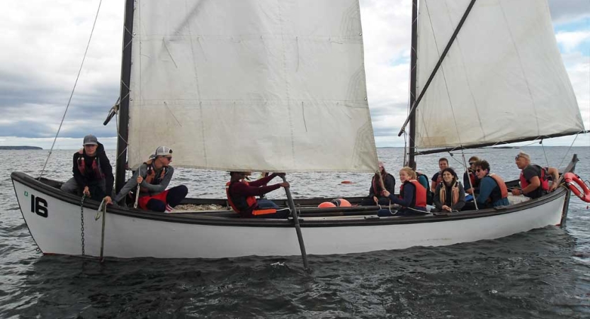 A group of people wearing life jackets sit in a sailboat with white sails. 